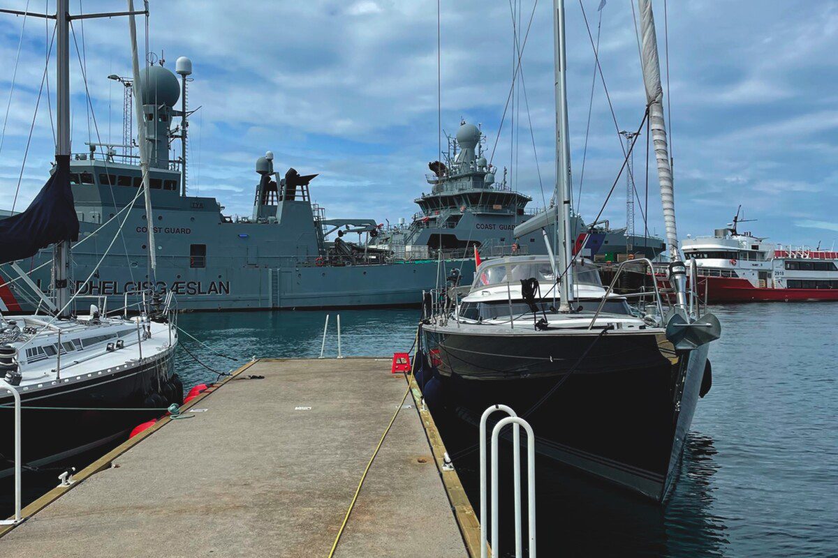 Large sailing yacht tied up to a dock with a grey warship in the back ground