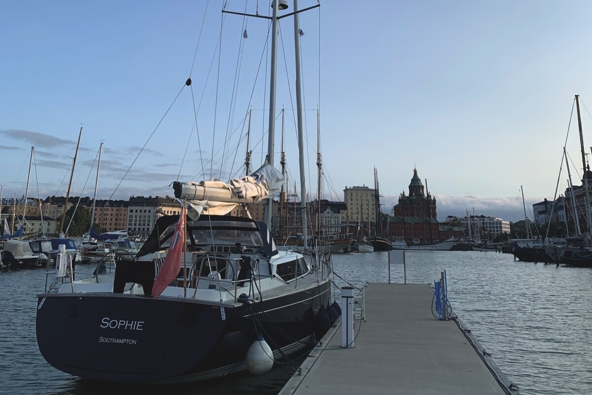 A blue hulled sailing yacht tied up alongside a dock with a cityscape in the distance